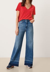 Rød V-hals t-shirt parret med brede blå jeans med en flosset kant. Modellen har blå sneakers på med røde og hvide detaljer.