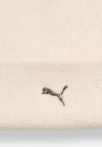 Bonnet en tricot crème avec une texture douce, ornée d'un logo Puma argent métallique sur le devant, offrant un contraste subtil avec le tissu.