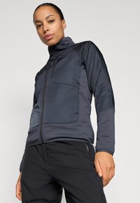 Veste zippée gris foncé avec un col haut, présentant des panneaux de tissu mats et brillants en contraste, ainsi qu'une poche sur la poitrine. Texture lisse.