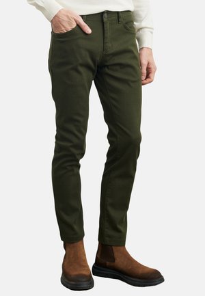 Mand iført olivengrønne slim-fit bukser med hånden i lommen og brune ruskind Chelsea-støvler på en hvid baggrund.