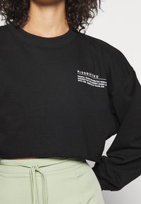 Sudadera negra corta con mangas largas, tejido texturizado y dobladillo sin acabados. Presenta una marca impresa en blanco en la parte delantera. Combinada con pantalones verde claro.