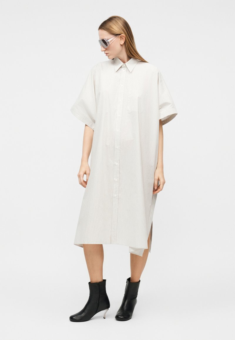 Femme portant une robe-chemise blanche oversize boutonnée, des bottines noires et des lunettes de soleil oversized, debout devant un fond uni.