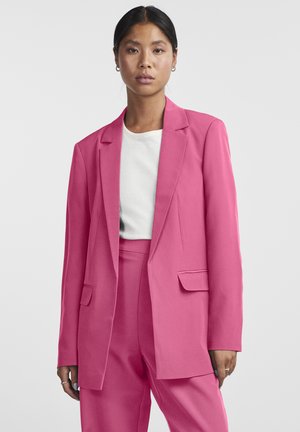 Blazer rosa acceso su misura con chiusura a un bottone, rever a incavo e due tasche frontali, abbinato a una maglia bianca a coste e pantaloni coordinati.
