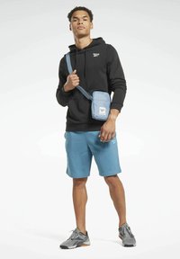 Bolso cruzado azul con una superficie texturizada, correa ajustable y un bolsillo delantero. El modelo lleva una sudadera negra y pantalones cortos azules.