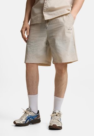 Mężczyzna ubrany w beżowe szorty ze sznurkiem, białe skarpetki i białe buty do biegania z niebieskimi i brązowymi detalami, stojący z jedną ręką w kieszeni.