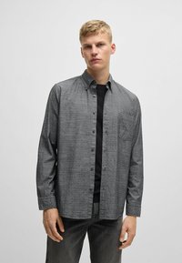 Camisa de botones en tela de color gris oscuro con una textura sutil, de mangas largas, con cuello abotonado y un bolsillo en el pecho. Llevada sobre una camiseta negra.