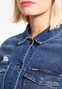 Sininen farkkupaita, jossa on klassinen kaulus, kaksi etutaskua ja näkyvät ompeluyksityiskohdat. Sisältää kuluneen alueen kankaassa.