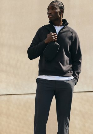 Ung mand med fletninger iført en sort sweatshirt og bukser, bærende en lille sort skuldertaske, stående foran en beige væg.