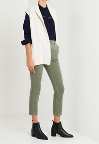 Elfenbensfärgad stickad väst över en marinblå skjorta med knappar, kombinerad med olivgröna byxor som når till anklarna och svarta läderstövlar som går upp till anklarna.