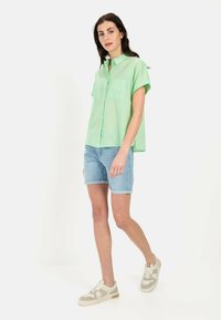 Hellgrünes kurzärmliges Button-down-Hemd mit zwei vorderen Taschen, kombiniert mit Denim-Shorts mit umgeschlagenem Saum und beigen Sneakers.