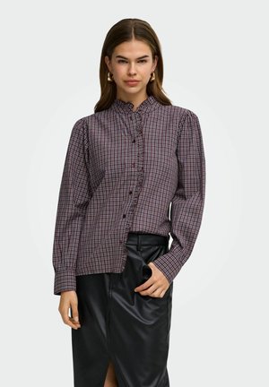 Jeune femme portant un chemisier à carreaux violet à manches longues avec un col à volants et une jupe en cuir noire, debout devant un fond uni.