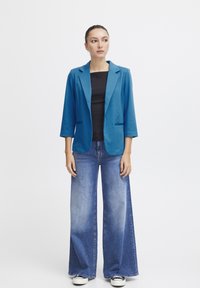 Tyrkysový blazer s třičtvrtinovými rukávy, spárovaný s volnými, světle modrými džínami. Modelka má na sobě černý top a bílé tenisky.