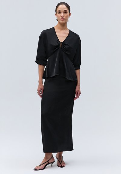 Femme portant un chemisier noir à manches longues avec col en V et détail lien, une longue jupe noire assortie et des sandales à brides à petit talon.