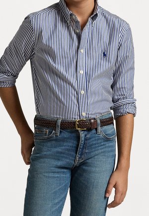 Niño con camisa de botones a rayas verticales azules y blancas con las mangas remangadas, cinturón trenzado marrón y vaqueros azules contra un fondo liso.