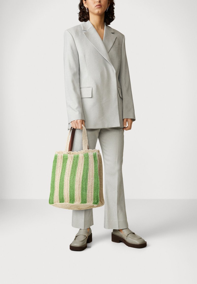 Femme en tailleur gris clair tenant un sac fourre-tout tissé beige et vert rayé, portant des mocassins compensés beige, debout devant un fond blanc.