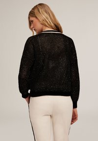 Maglione di rete nera con orlo e polsini a costine, caratterizzato da una vestibilità ampia. Indossato con pantaloni color crema, che mostrano un motivo testurizzato.