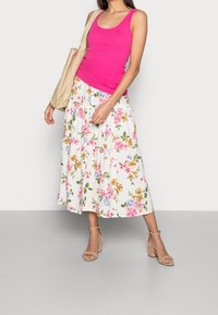Top de tirantes ajustado rosa combinado con una falda blanca fluida que presenta un patrón floral colorido. La modelo lleva un bolso tote beige y calza tacones nude.