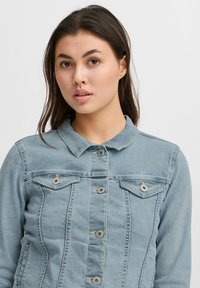 Chaqueta de mezclilla azul clara con cierre de botones, dos bolsillos en el pecho, cuello puntiagudo y detalles de costura en la parte delantera y los lados.