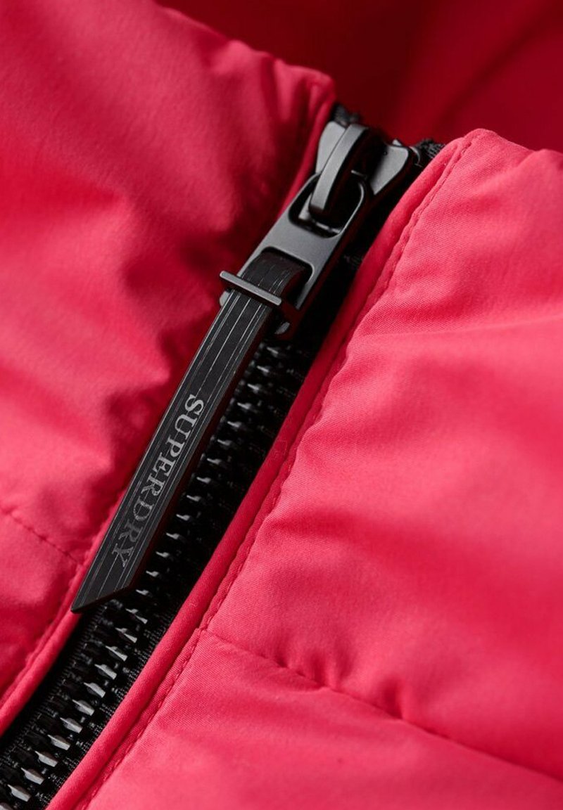 Roter gestepptes Gewebe mit einem schwarzen Reißverschluss. Der Reißverschluss hat einen strukturierten Zugtab, der mit "SUPERDRY" versehen ist. Nahaufnahme von Naht und Materialstruktur.