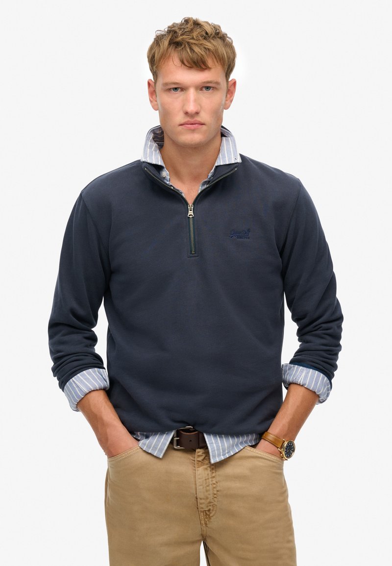 Suéter navy con cremallera y una camisa de cuello azul claro debajo, combinado con pantalones caqui. Presenta puños acanalados y detalle del logo.