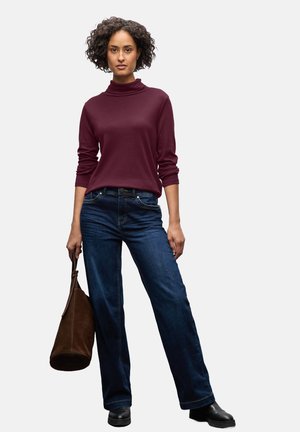 Burgundy turtleneck, relaxed fit, paired with dark blue wide-leg jeans. Model holds a brown suede handbag, wearing black footwear.