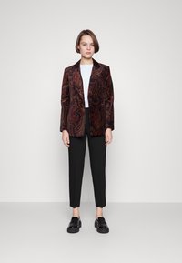 Blazer en velours bordeaux avec motif paisley, associé à un haut blanc et à un pantalon noir fuselé ; mannequin portant des chaussures à plateforme noires.