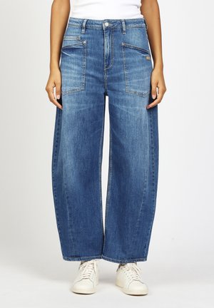 Frau trägt weit geschnittene blaue Jeans mit aufgesetzten vorderen Taschen, weiße Sneakers und ein weißes Oberteil, steht vor einem einfarbigen Hintergrund.