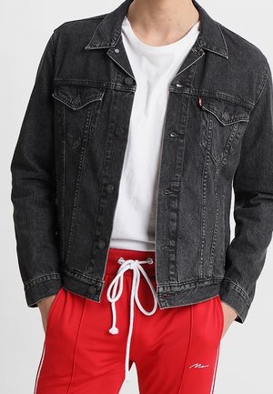 Person iført en sort denimjakke over en hvid t-shirt og røde træningsbukser med hvide snore og sidelommer.