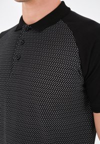 Polo noir avec un devant à motifs avec des lignes blanches en zigzag, des manches courtes et un col noir uni à trois boutons.