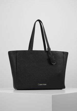 Czarna torba typu tote Calvin Klein z dwoma paskami na ramię oraz małą zawieszką, umieszczona na białym postumencie na szarym tle.