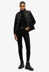 Chaleco acolchado negro con cuello alto, cierre de cremallera y logotipo, llevado sobre un suéter de cuello alto negro y vaqueros ajustados negros, combinado con botas de combate negras.