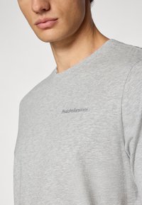 Graues Baumwoll-T-Shirt mit Rundhalsausschnitt, das ein kleines gesticktes "Peak Performance"-Logo auf der Brust trägt. Glatte Textur.