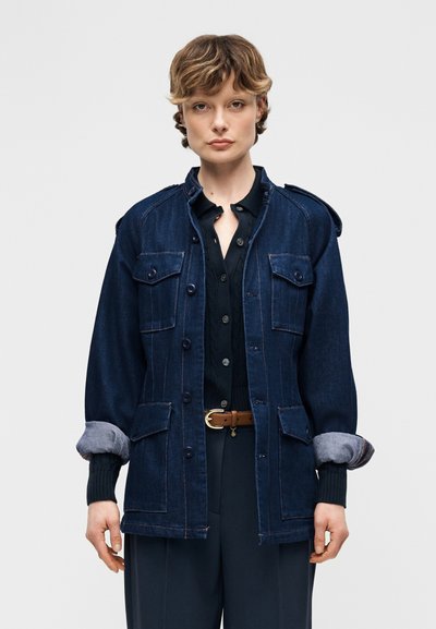 Άτομο που φοράει ένα σκούρο μπλε τζιν μπουφάν με γυρισμένα μανίκια πάνω από ένα μαύρο πουκάμισο με κουμπιά και ναυτικά μπλε παντελόνια με καφέ ζώνη.