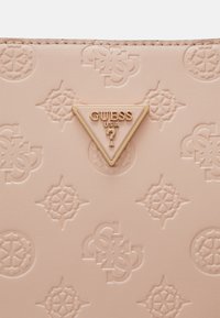 Vaaleanpunainen kohokuvioitu käsilaukku, jossa on geometrinen kuvio, kultainen kolmionmuotoinen logo "GUESS USA", sileä pinta ja hienovarainen kiilto.