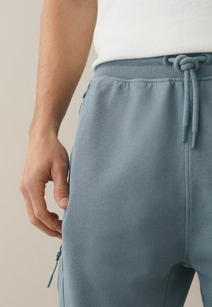 Lähivõte inimesest, kes kannab helesiniseid dressipükse nööriga vöökohal ja külje lukuga taskuga, seistes vasak käsi lõdvestunult.