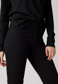 Vaqueros pitillo negros con una textura suave, cintura alta y detalles de costura sutiles. Combinados con un suéter negro de manga larga.