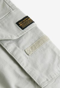 Lichtgrijze stofpatch met een zwart en gouden label met de tekst "NEXT SUPPLY." Bevat daarnaast een gestructureerde beige banddetails.