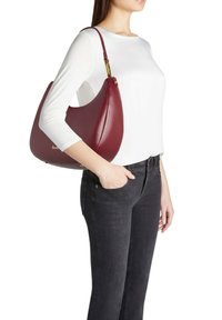 Borsa hobo in pelle color bordeaux con forma curva, dettagli dorati e una texture liscia. Indossata a spalla da una persona con una maglietta bianca.