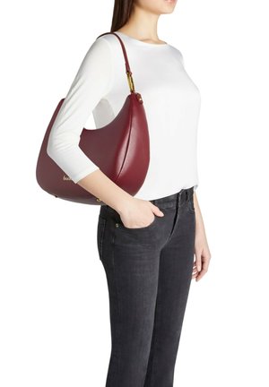 Borsa hobo in pelle color bordeaux con forma curva, dettagli dorati e una texture liscia. Indossata a spalla da una persona con una maglietta bianca.