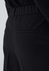 Gros plan sur un pantalon noir sur mesure avec une poche arrière passepoilée et une ceinture cousue, porté avec un haut noir.