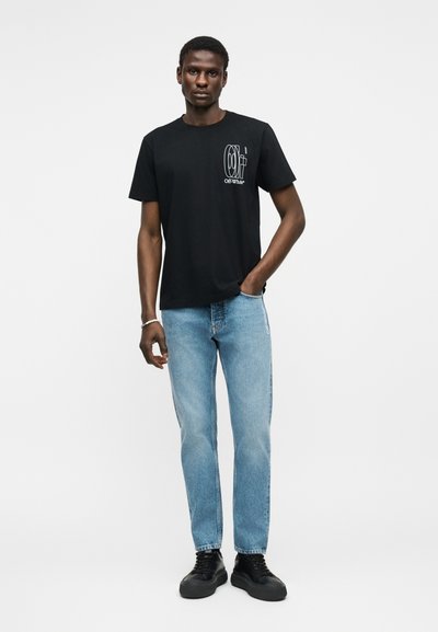 Άνδρας που στέκεται με το ένα χέρι στην τσέπη, φορώντας μαύρο μπλουζάκι Off-White με γραφικά, ανοιχτό μπλε τζιν και μαύρα αθλητικά παπούτσια σε λευκό φόντο.