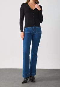 Mujer vistiendo un cárdigan negro con botones, jeans azules de cintura alta con bolsillos frontales y botas negras de tacón, de pie contra un fondo liso.
