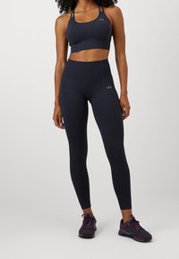 Ciemnoniebieski stanik sportowy i legginsy z wysokim stanem w prążkowanej tkaninie, w połączeniu z fioletowymi butami sportowymi. Posiada minimalne logo i regulowane ramiączka.