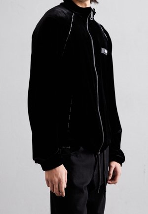 Persona con una chaqueta negra con cremallera y cuello alto, con un pequeño parche en el pecho, de pie frente a un fondo claro y uniforme.