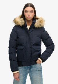 Veste matelassée bleu marine avec une capuche ornée de fourrure, deux poches avant et des poignets côtelés ; elle possède une fermeture éclair et une poche sur le côté.