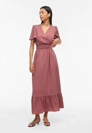 Kvinne i lilla omslagskjole med puffermene, rynket midje, volangkant og svarte, åpne hælsandaler, stående mot en hvit bakgrunn.