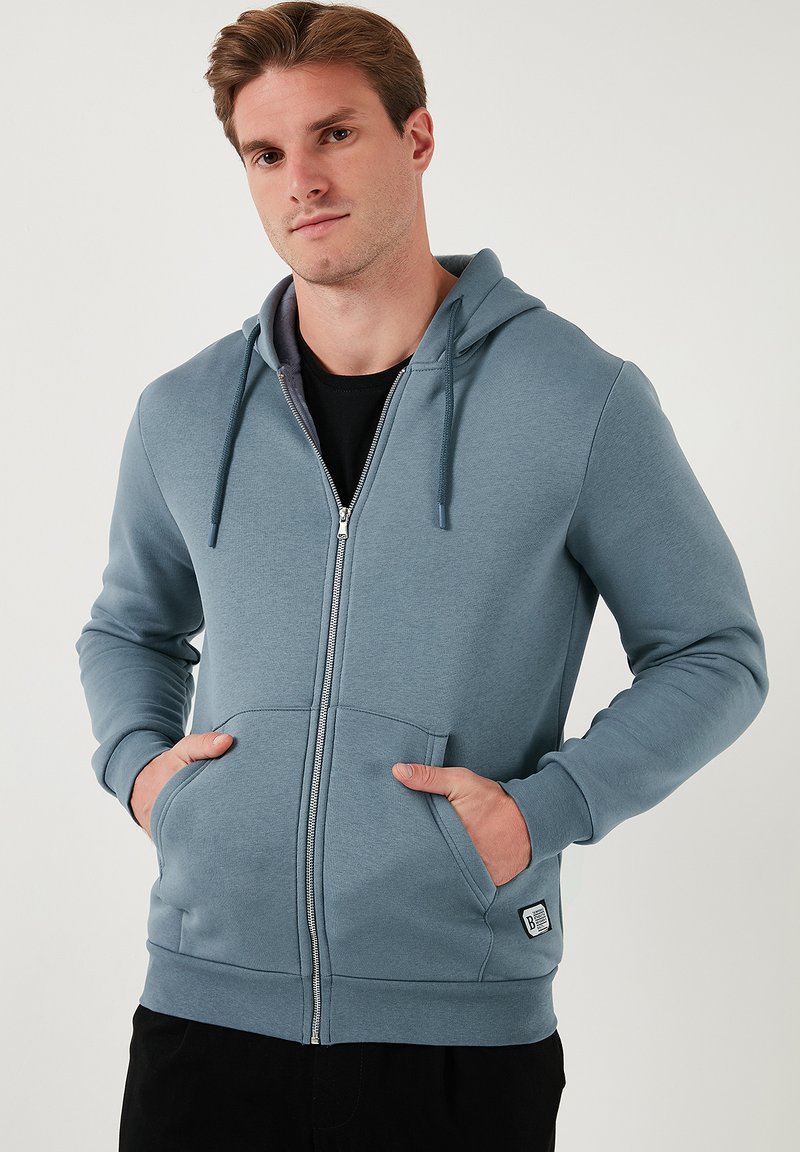 Sudadera con cremallera de color azul claro, hecha de una tela suave. Cuenta con una capucha ajustable, bolsillos frontales y puños acanalados, además de una etiqueta con el logo en la parte inferior.
