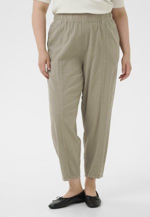 Femme portant un pantalon beige à taille élastique associé à des ballerines noires, debout devant un fond blanc uni.