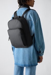Schwarzer Rucksack mit glatter, matter Oberfläche, rechteckiger Form, Fronttasche und gepolsterten Schulterriemen. Über einer hellblauen Jeansjacke getragen.