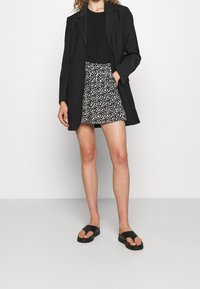 Zwarte blazer over een zwart topje, gecombineerd met een korte wit-zwarte rok met patroon en zwarte schuifzandalen.